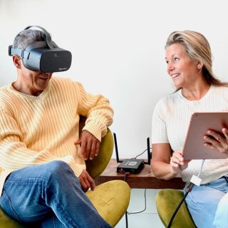 A man wearing a Vision Buddy headset sits on a green chair while a smiling woman next to him holds a tablet and interacts with him.