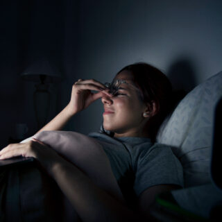 A tired woman in bed rubbing her eyes while wearing glasses, with a laptop on her lap and a clock showing late night hours.