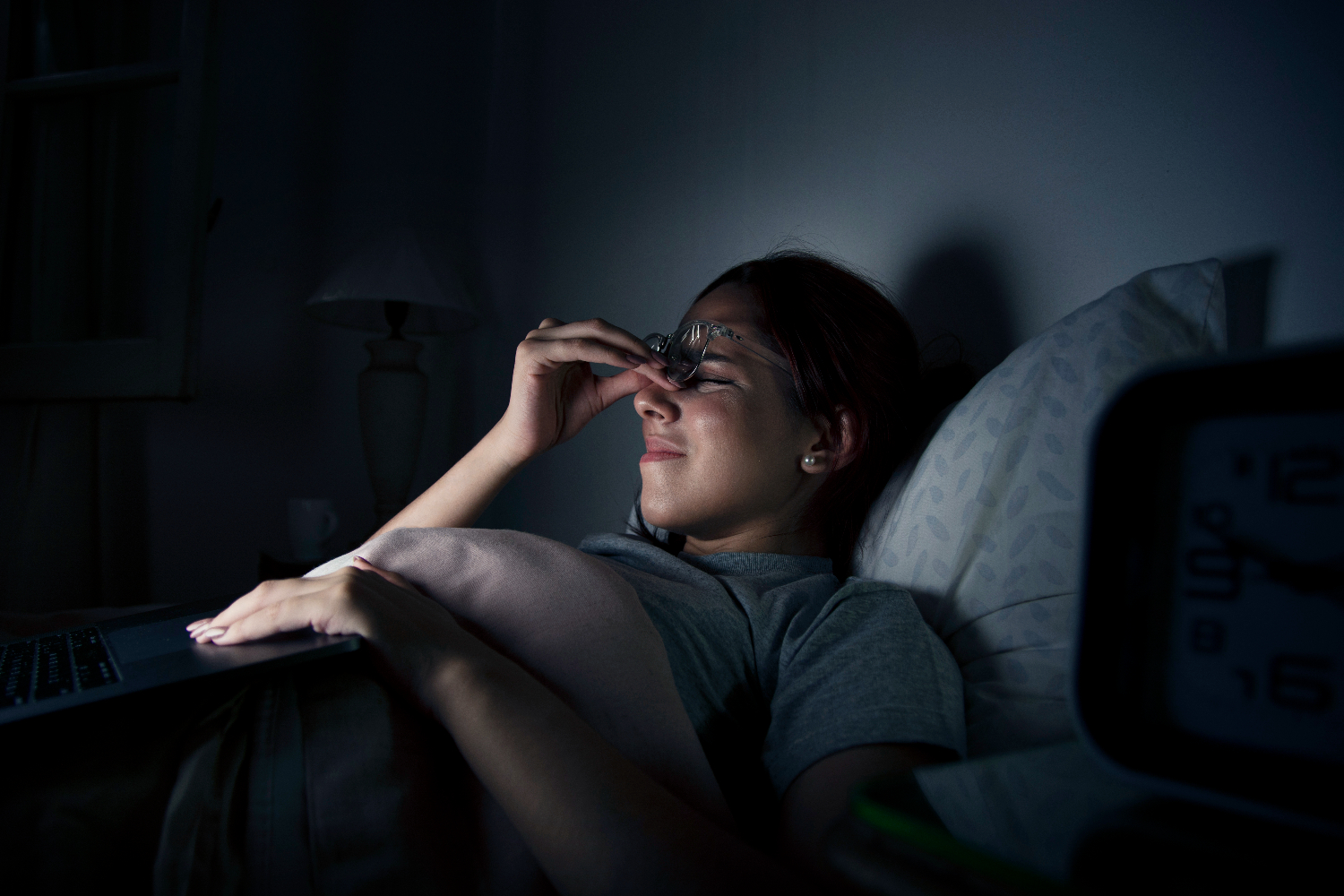 A tired woman in bed rubbing her eyes while wearing glasses, with a laptop on her lap and a clock showing late night hours.