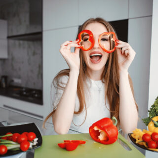 A smiling woman in a kitchen holds red bell pepper slices like glasses, surrounded by fresh vegetables on a cutting board.