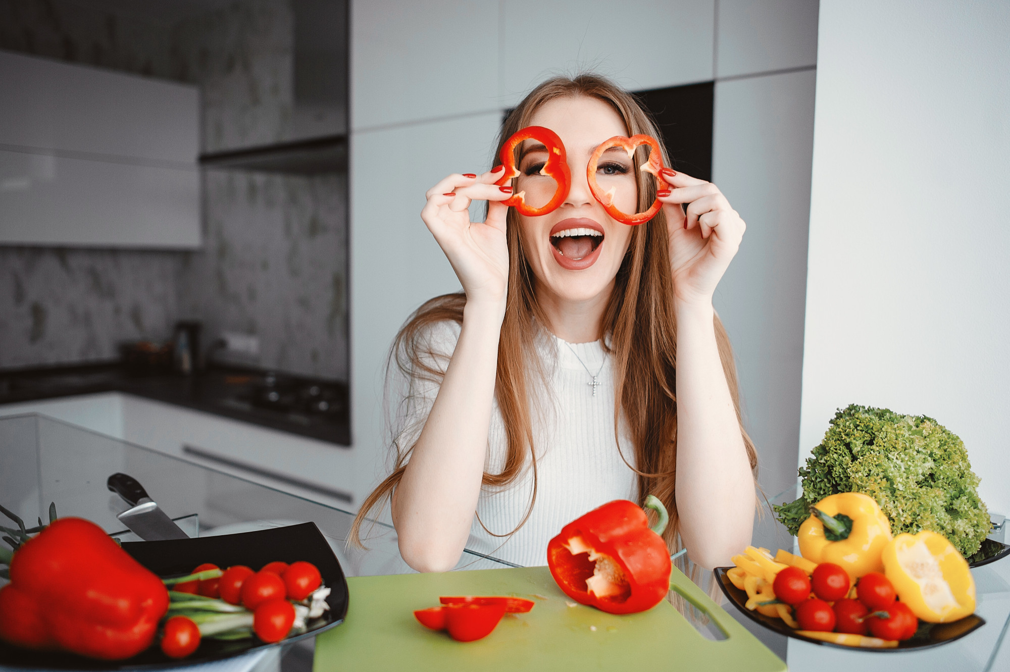 A smiling woman in a kitchen holds red bell pepper slices like glasses, surrounded by fresh vegetables on a cutting board.