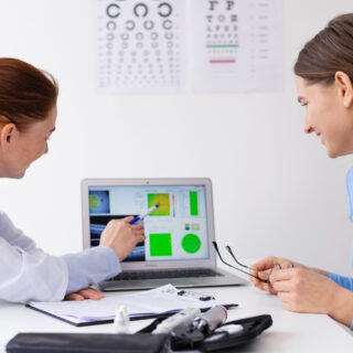 An eye doctor explains test results to a smiling patient using a laptop, with eye charts visible on the wall behind them.