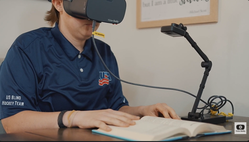 Caleb, wearing a U.S. Blind Hockey Team jersey, reading a book with his Vision Buddy headset and CCTV Mini